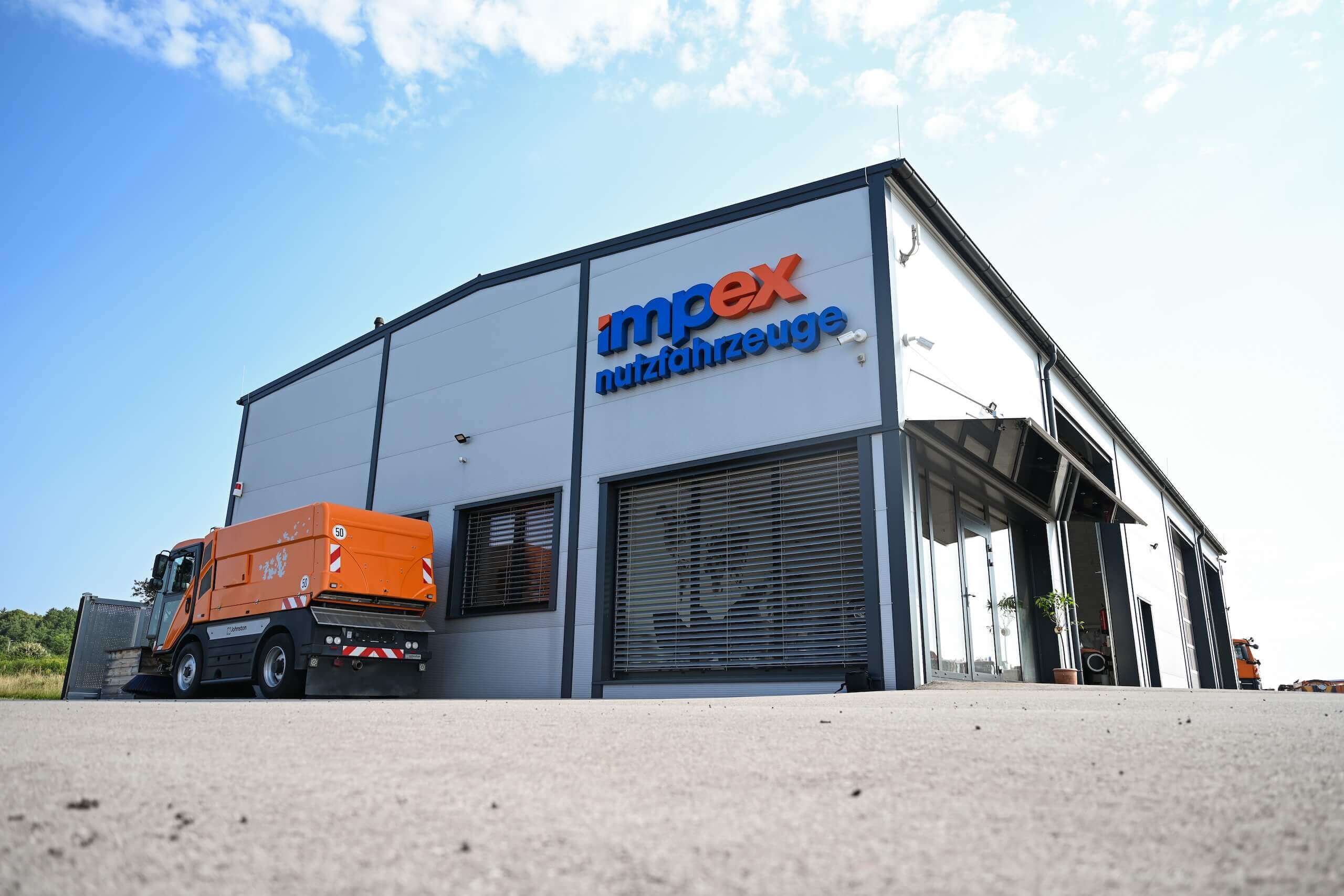 Lagerhalle von Impex Nutzfahrzeuge mit orangefarbenem LKW im Vordergrund und blauem Himmel im Hintergrund.
